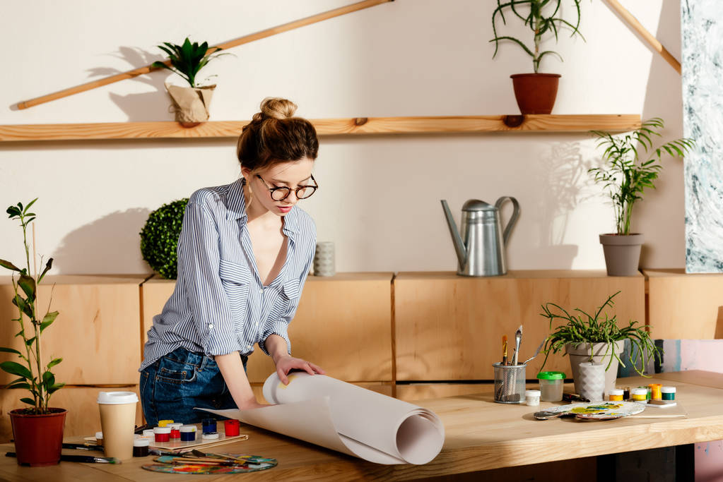 attraktive stylische Frau mit Brille rollt Leinwand auf Tisch mit Malzubehör  - Foto, Bild