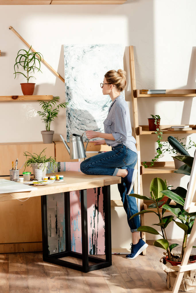 side view of stylish female artist in eyeglasses watering potted plan in studio - Photo, Image