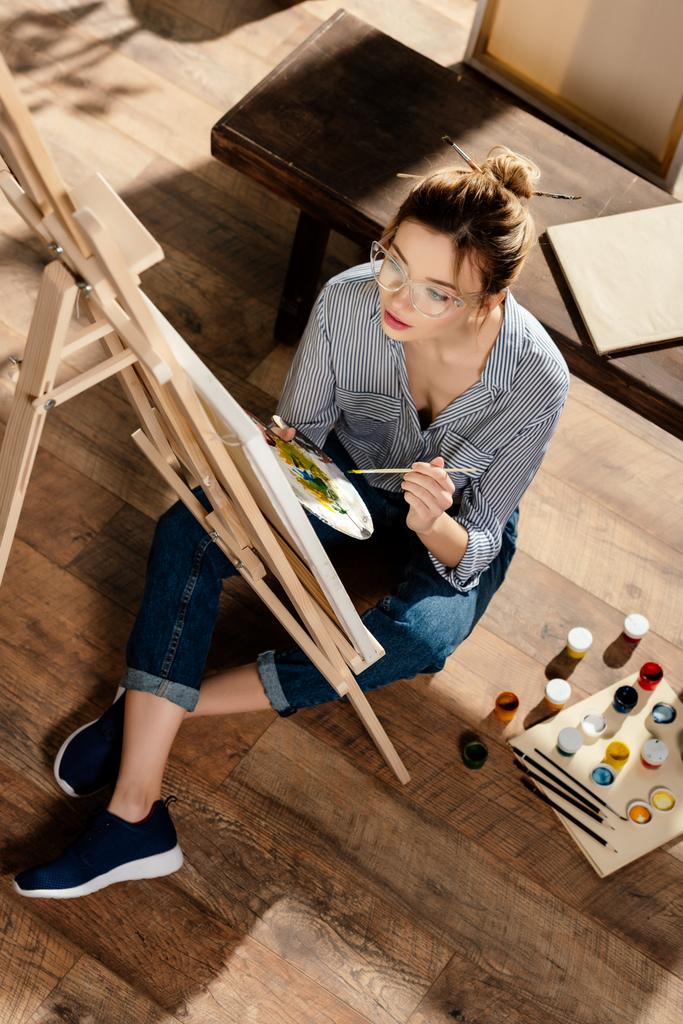 Hochwinkelaufnahme einer stilvollen Künstlerin in Brillenmalerei auf Staffelei - Foto, Bild