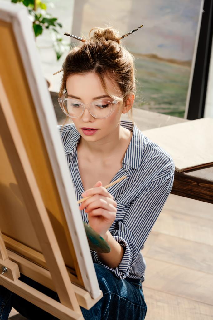 イーゼルの絵画の眼鏡で若いおしゃれな女性アーティスト  - 写真・画像