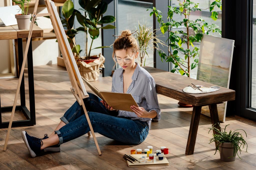 стильная молодая художница в очках читает книгу и сидит на полу с принадлежностями для живописи
 - Фото, изображение