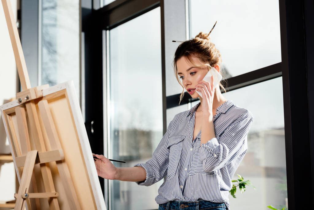jovem artista feminina falando no smartphone e pintura no cavalete
  - Foto, Imagem