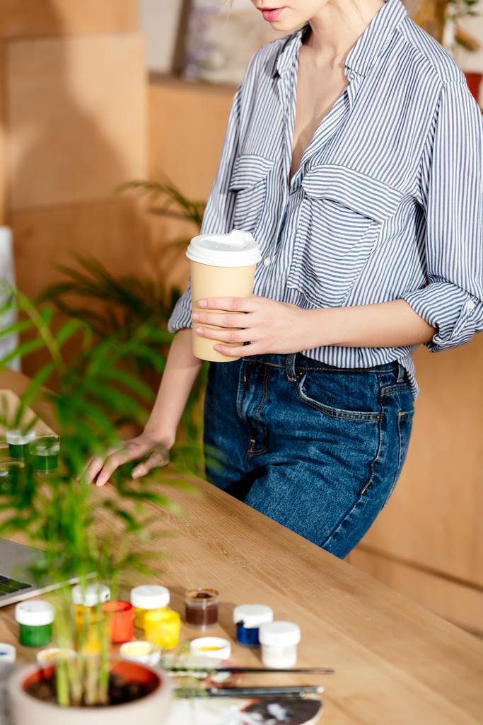 обрізаний знімок молодої жінки, що тримає паперову чашку кави за столом з ноутбуком і фарбами
 - Фото, зображення