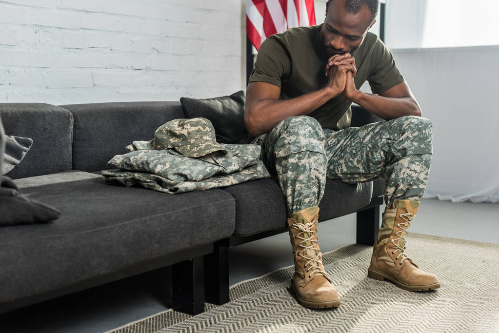 Nachdenklicher Soldat sitzt auf Sofa und betrachtet seine Tarnkleidung - Foto, Bild