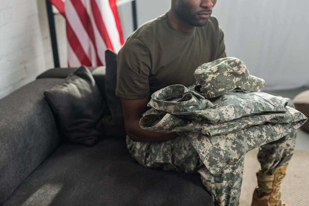 Bonito soldado segurando suas roupas de camuflagem enquanto sentado no sofá
 - Foto, Imagem