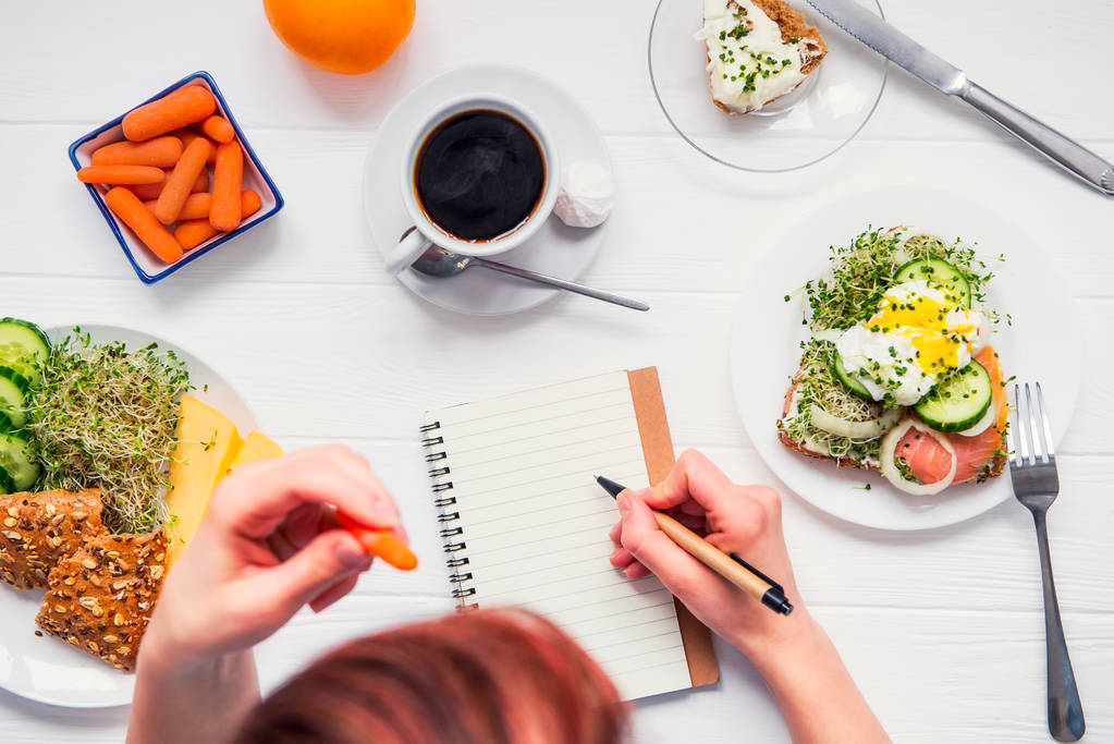 成功した人々 の朝の習慣。日の計画、健康な食事。女性はニンジンを食べると、先着でノートに書いて朝食白い木製のテーブルのため。トップ ビューの選択と集中. - 写真・画像