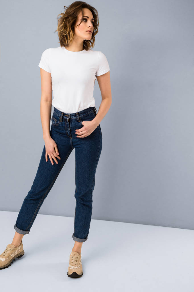 Portret stylowy młoda dziewczyna w podstawowe biały t-shirt i wysokiej wcięty jeans niebieski pozowanie Studio na szarym tle - Zdjęcie, obraz