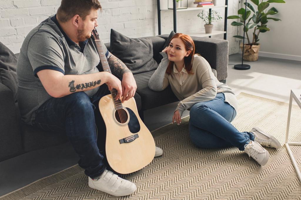 size plus boyfriend holding acoustic guitar and looking at girlfriend at home - Photo, Image