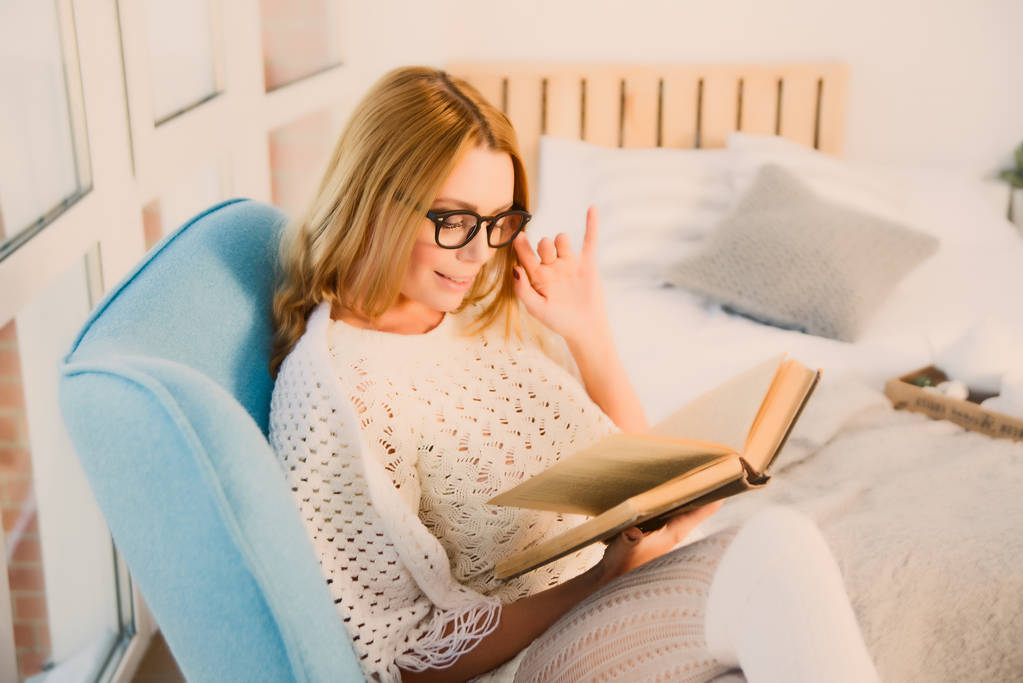 Jonge blonde vrouw thuis zit op moderne stoel voor venster ontspannen in haar woonkamer lezen boek  - Foto, afbeelding