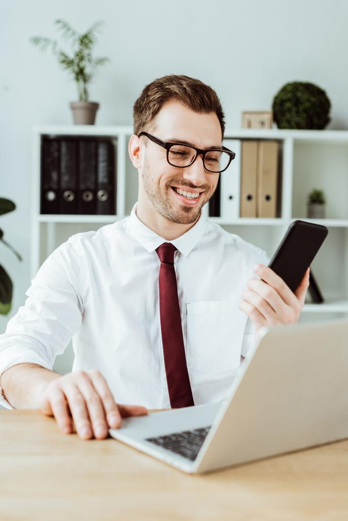 komea liikemies, joka käyttää älypuhelinta ja kannettavaa tietokonetta työpaikalla nykyaikaisessa toimistossa
 - Valokuva, kuva