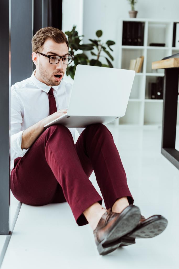 shocked businessman in eyeglasses using laptop while sitting on floor in office - Photo, Image