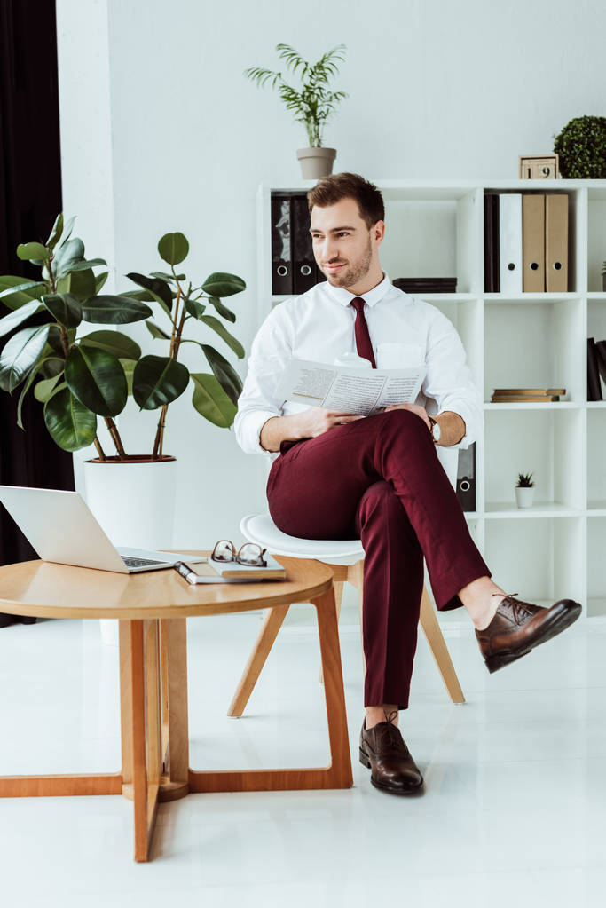 homem de negócios elegante bonito com jornal e laptop sentado no escritório moderno
 - Foto, Imagem