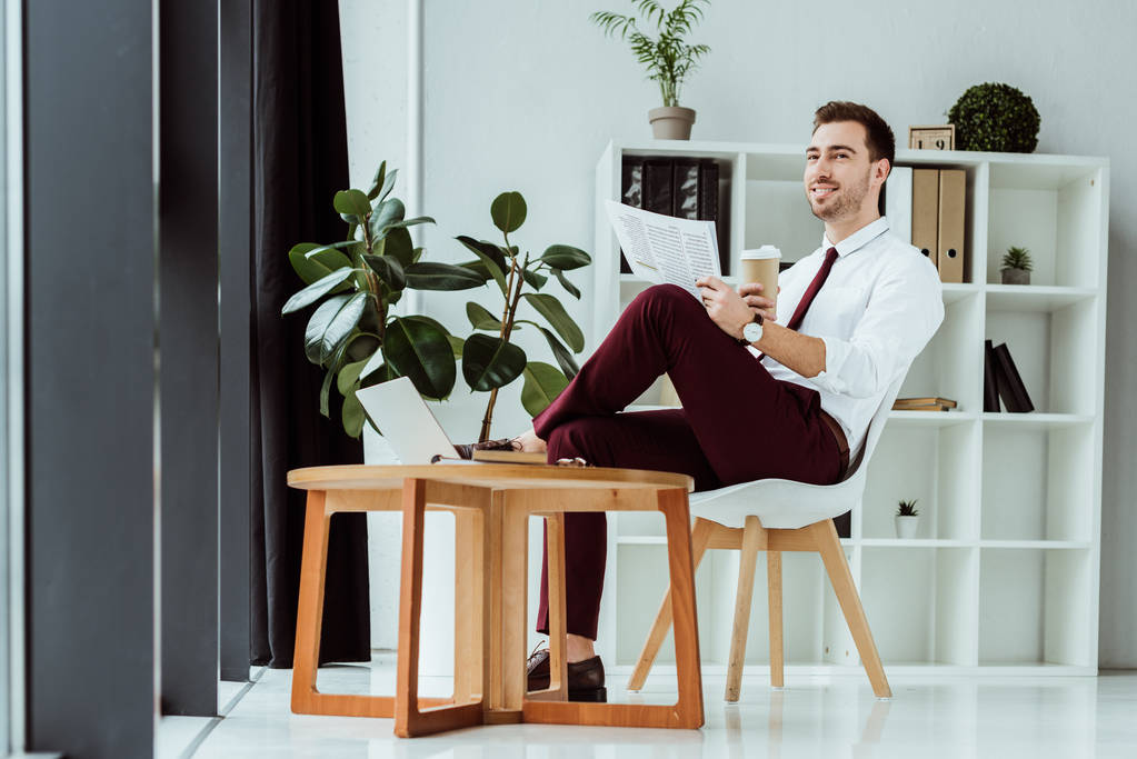 stylish businessman with coffee reading newspaper in modern office with laptop - Photo, Image