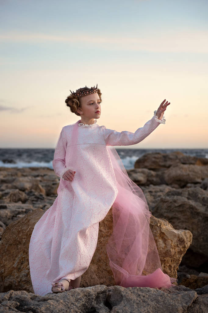 caprichosa menina princesa inquieta sentado em uma pedra junto ao mar em um vestido rosa com véu e coroa
 - Foto, Imagem