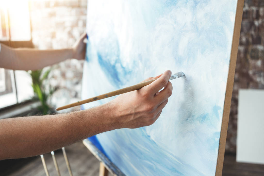 Чоловіча рука художника крупним планом тримає пензлик на тлі полотна на мольберті. Художник малює океанічну хвилю в студії лофт. Ефект спалаху. концепція хобі
 - Фото, зображення
