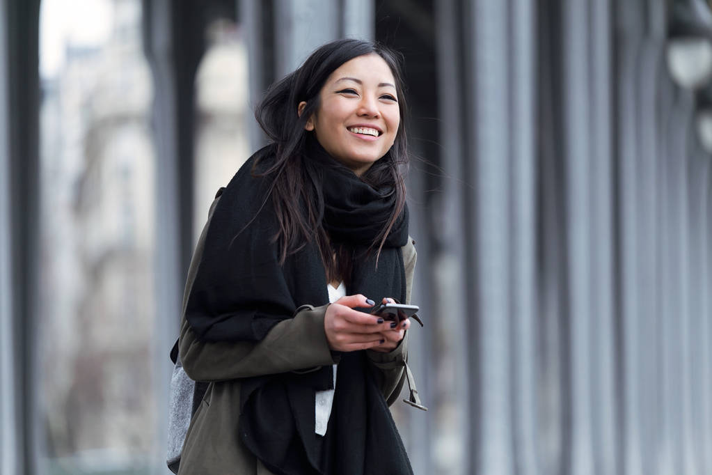 Улыбающаяся азиатская молодая женщина, использующая свой мобильный телефон на улице
. - Фото, изображение