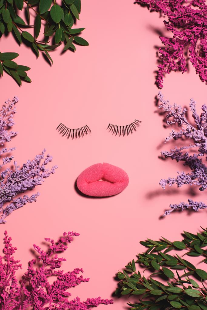 vista superior da face feita de esponja e pestanas falsas cercadas de flores na superfície rosa
 - Foto, Imagem