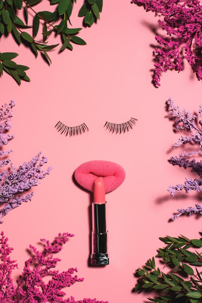 vista superior de la cara hecha de esponja con pestañas postizas y lápiz labial rodeada de flores en la superficie rosa
 - Foto, Imagen