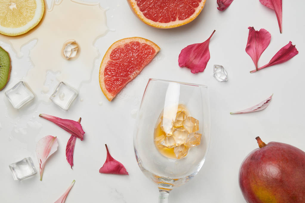 白い表面上のワイングラス、エキゾチックなフルーツ、アイス キューブ、花びらの配置平面図 - 写真・画像