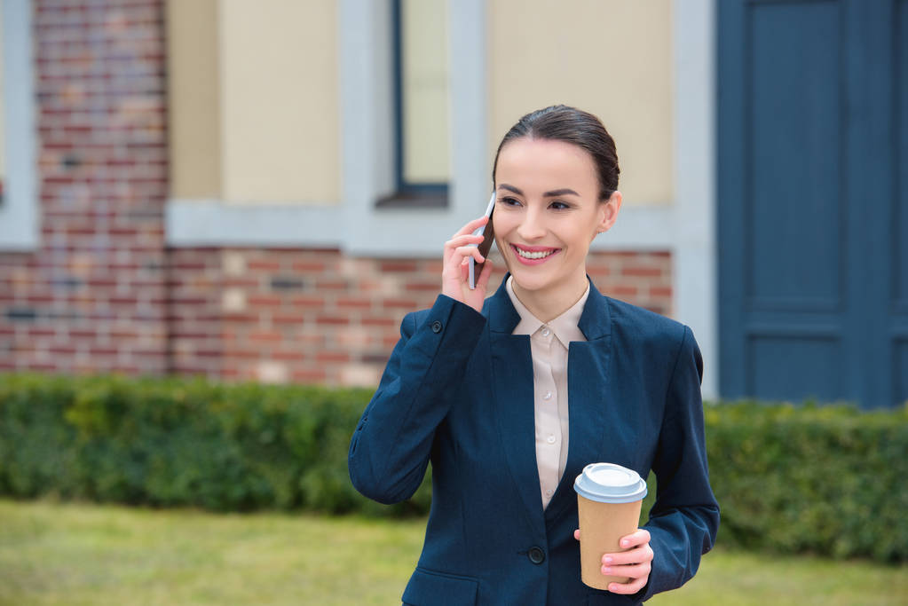 Schöne Geschäftsfrau spricht per Smartphone und hält Einweg-Kaffeetasse in der Hand - Foto, Bild