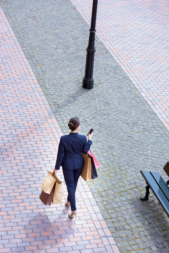 Blick aus der Vogelperspektive auf Geschäftsfrau, die mit Einkaufstaschen spaziert und das Smartphone benutzt - Foto, Bild