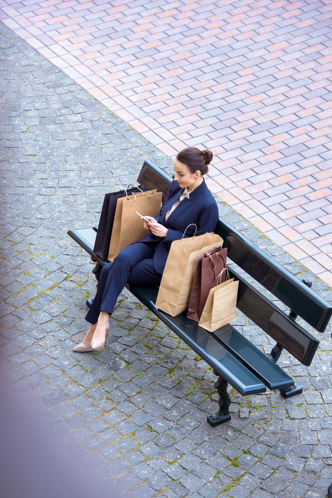 высокий угол зрения привлекательных деловых женщин, сидящих с мешками для покупок на скамейке и с помощью смартфона
 - Фото, изображение