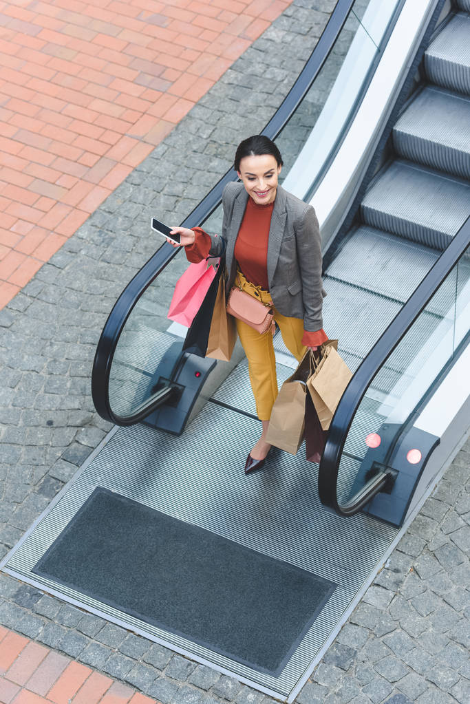 Blick von oben auf attraktive Frau, die mit Einkaufstaschen von Rolltreppe in Einkaufszentrum geht - Foto, Bild