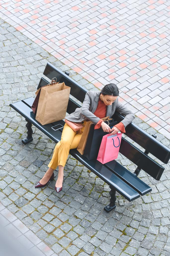 vista de ángulo alto de la mujer atractiva sentada en el banco y mirando en la bolsa de compras
 - Foto, imagen