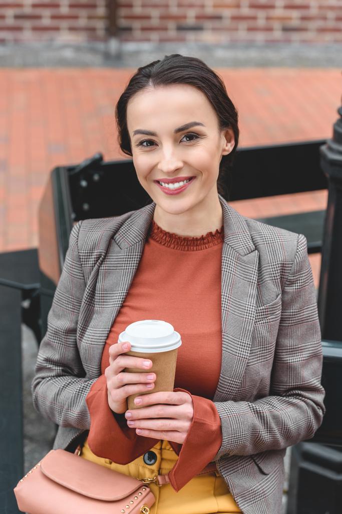 beautiful stylish woman holding coffee in paper cup and looking at camera - Photo, Image