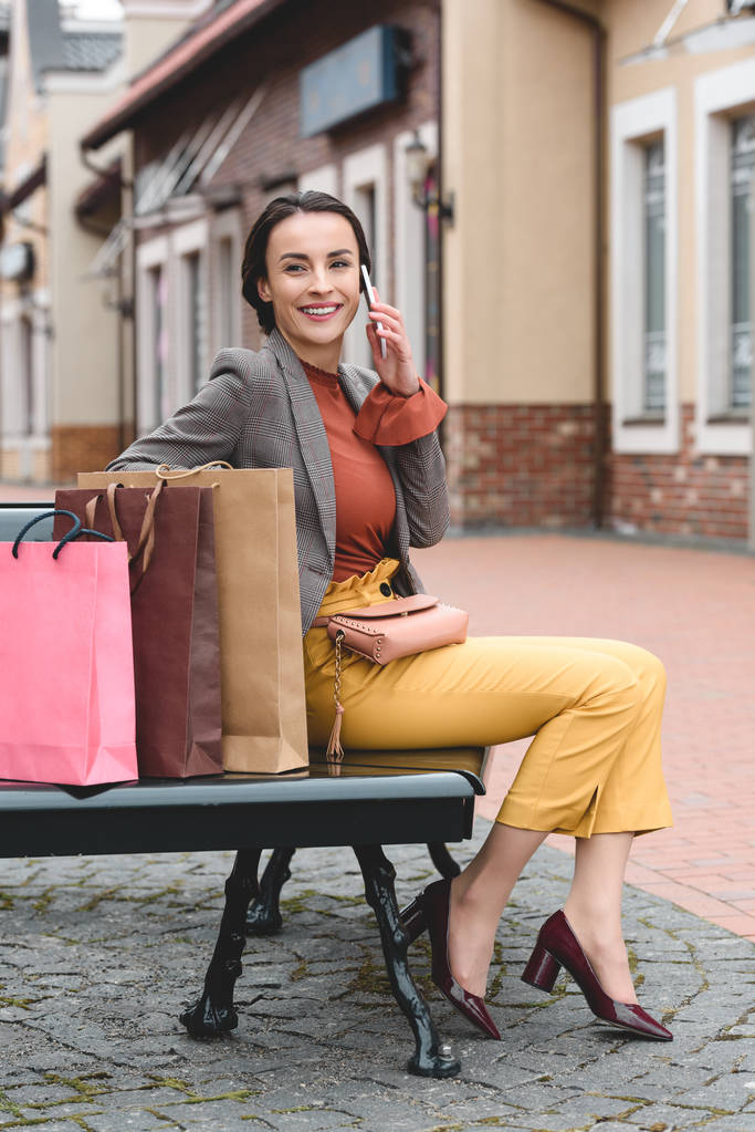όμορφη γυναίκα κάθεται σε παγκάκι με Τσάντες για ψώνια και να μιλάμε με smartphone - Φωτογραφία, εικόνα