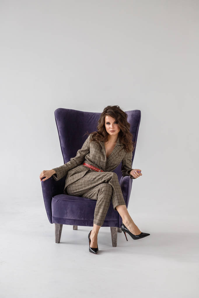 Retrato de estudio de moda de una hermosa mujer rizada que usa un traje casual a cuadros con cinturón rojo, sentado en un sillón púrpura, fondo de pared blanco
  - Foto, Imagen