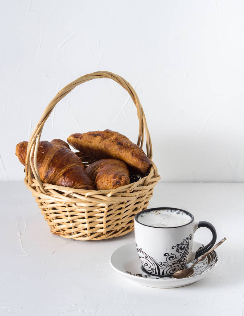 Morning breakfast: a mug of coffee with cream with fresh croissants in a basket. - Photo, Image