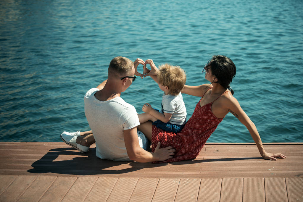 幸せな家族の時間、一緒に海の背景を過ごします。息子を持つ親は、リアビューで、海辺に座る。母と父の心や愛を作る彼らの子供に近い手でジェスチャーをします。家族での休暇の概念. - 写真・画像