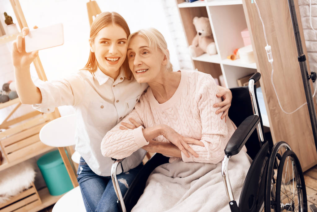 Κορίτσι φροντίδα ηλικιωμένης γυναίκας στην αναπηρική καρέκλα στο σπίτι και λήψη selfie σε smartphone  - Φωτογραφία, εικόνα