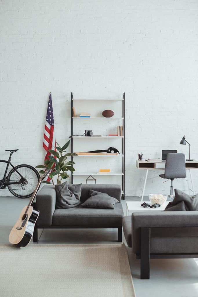 Interieur eines modernen Wohnzimmers mit Fahrrad, Gitarre, amerikanischer Flagge, Laptop, Regalen und Sesseln  - Foto, Bild