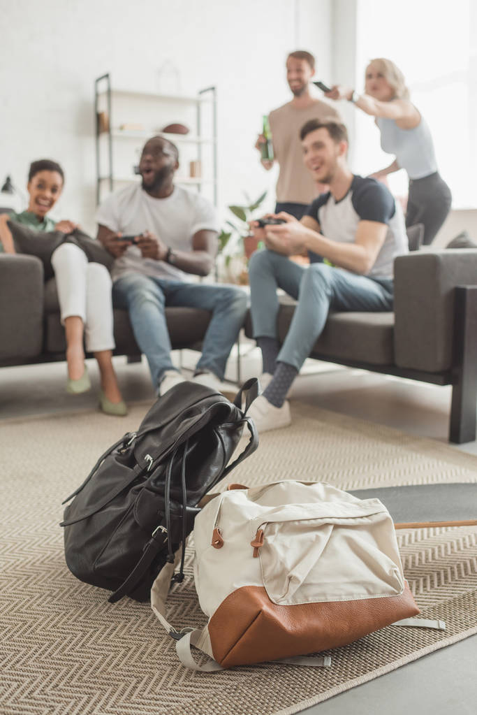  two backpacks and longboard on foreground and group of multiethnic friends with joysticks playing video game - Photo, Image