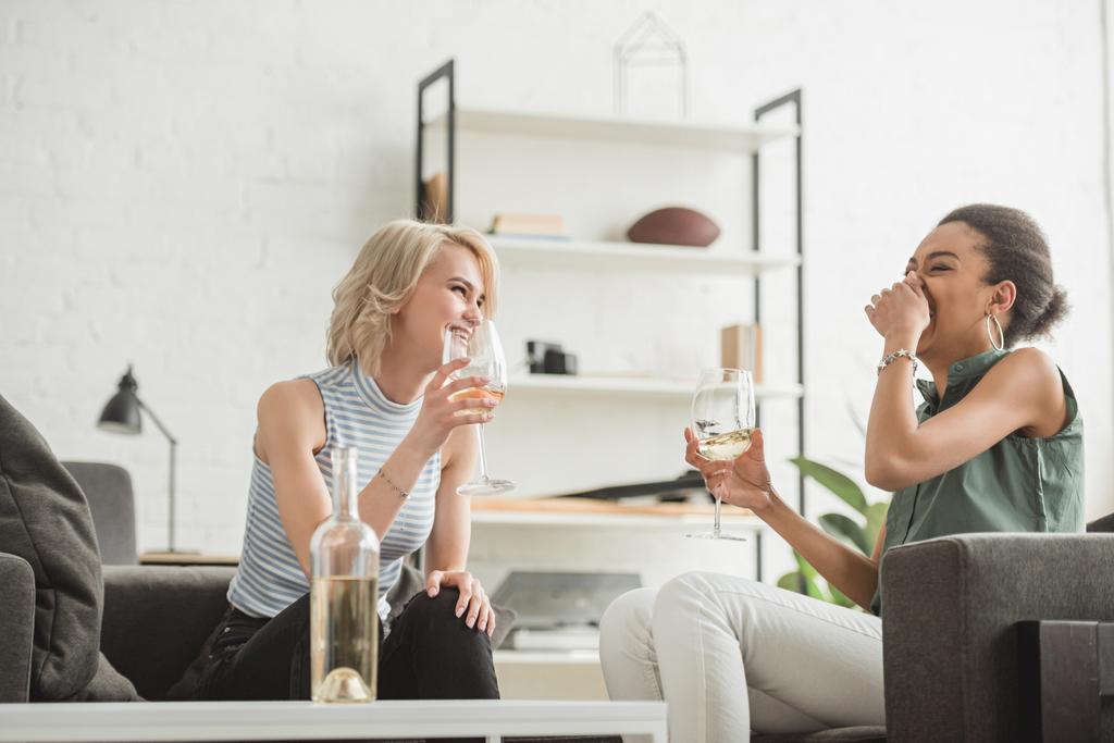 junge lachende multiethnische Freundinnen sitzen mit Gläsern Weißwein  - Foto, Bild