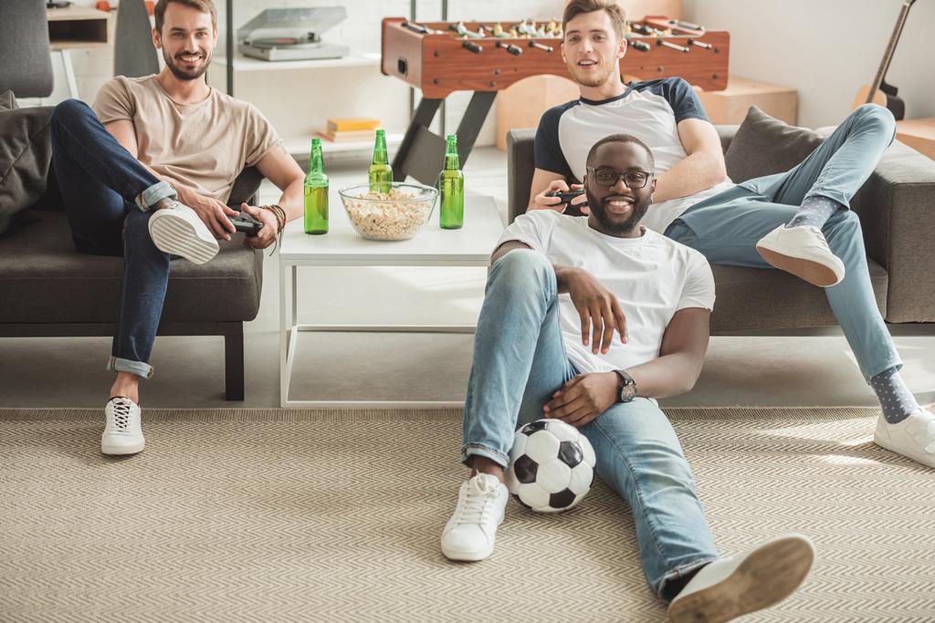 junger schwarzer Mann sitzt auf Teppich mit Ball zwischen zwei Freunden und spielt Videospiel mit Steuerknüppeln in der Hand - Foto, Bild