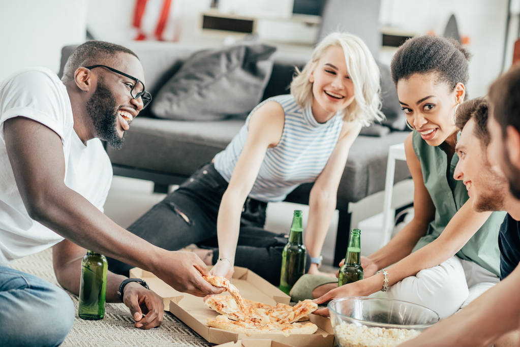 gente multiculturale sorridente seduta sul pavimento con pizza, popcorn e bottiglie di birra
  - Foto, immagini