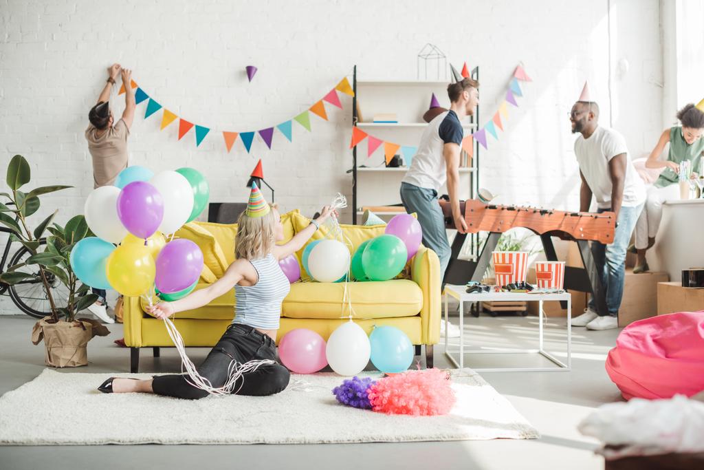 молодая женщина сидит на полу с красочными воздушными шарами и ее друзья украшают комнату позади
 - Фото, изображение