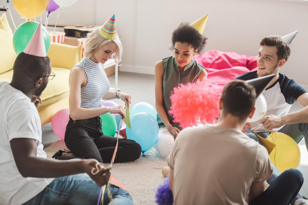 Gruppe junger multikultureller Freunde in Partyhüten sitzt mit Luftballons auf dem Boden im dekorierten Raum  - Foto, Bild