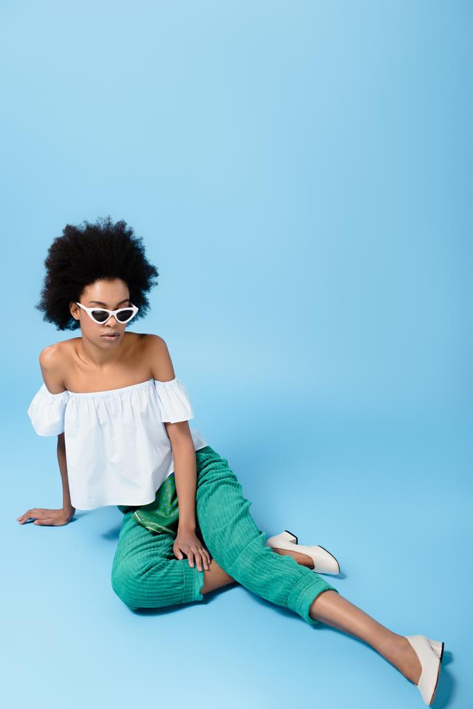 όμορφη νεαρή γυναίκα σε μοντέρνα ρούχα και τα γυαλιά ηλίου που κάθεται στο πάτωμα με μπλε - Φωτογραφία, εικόνα