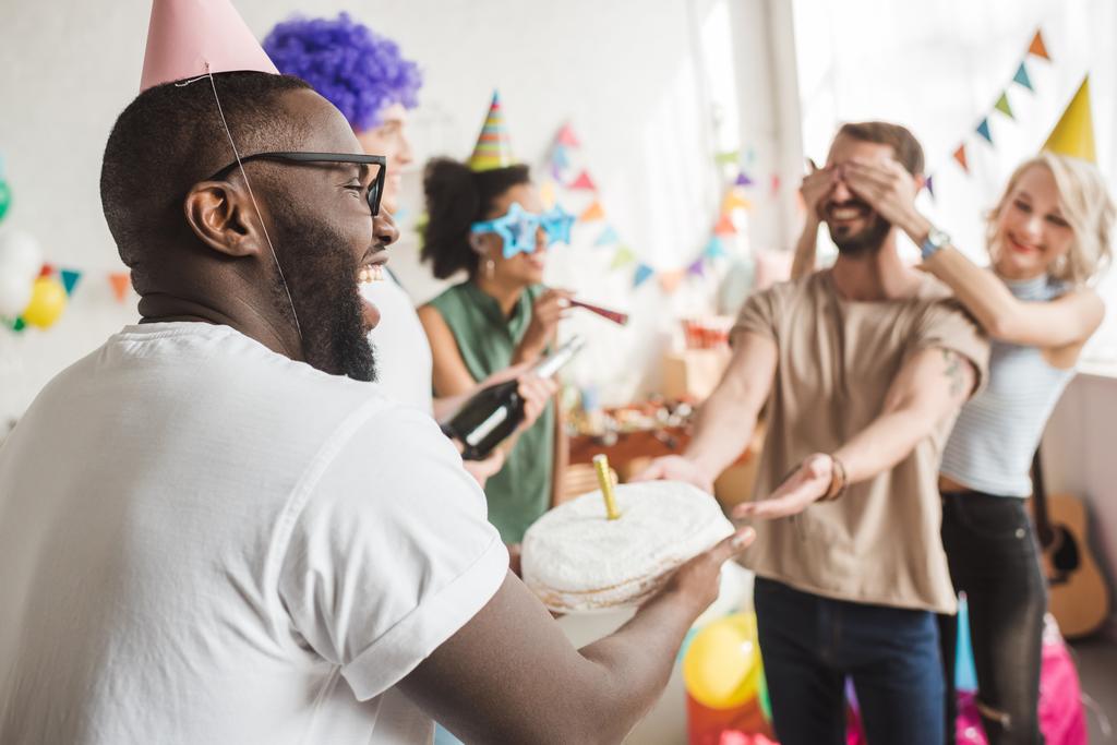Счастливые друзья, закрывая глаза молодому человеку и приветствуя его праздничным тортом
 - Фото, изображение