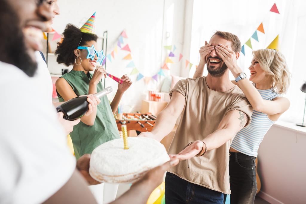 Усміхнені молоді люди прикривають очі молодого друга і вітають його з тортом на день народження
 - Фото, зображення