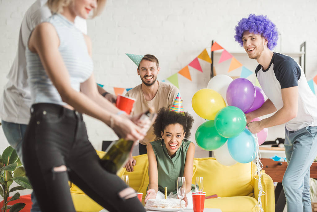 Jovens alegres olhando para a mulher abrindo champanhe na festa de aniversário
 - Foto, Imagem