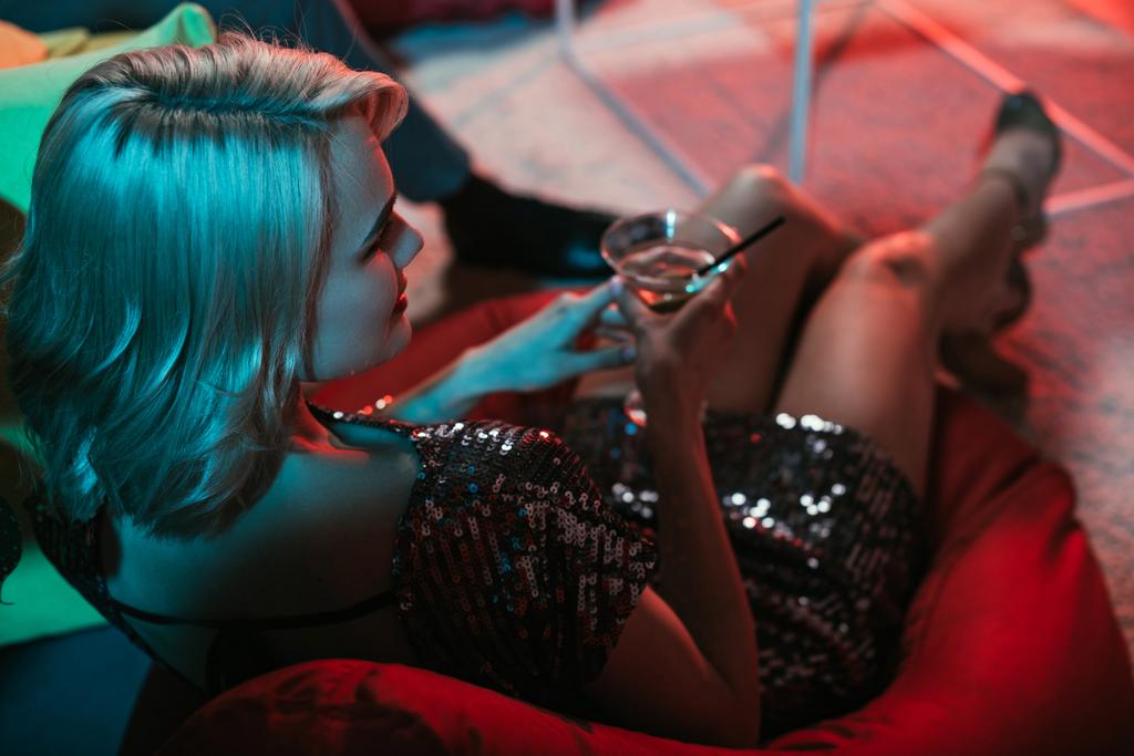 Blonde woman holding glass with cocktail while sitting in beanbag  - Photo, Image