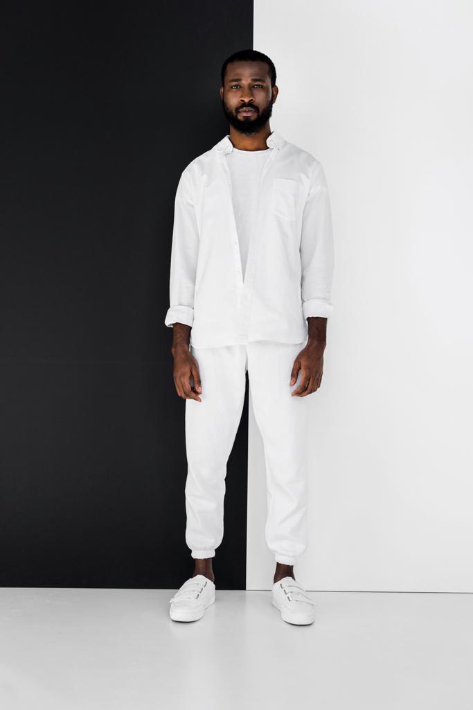 knappe stijlvolle Afro-Amerikaanse man in witte kleren in de buurt van zwart-witte muur - Foto, afbeelding