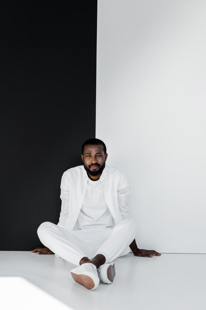 όμορφος μοντέρνος αφρικανική αμερικανική άνθρωπος που κάθεται στο πάτωμα κοντά σε μαύρο και άσπρο τοίχο - Φωτογραφία, εικόνα