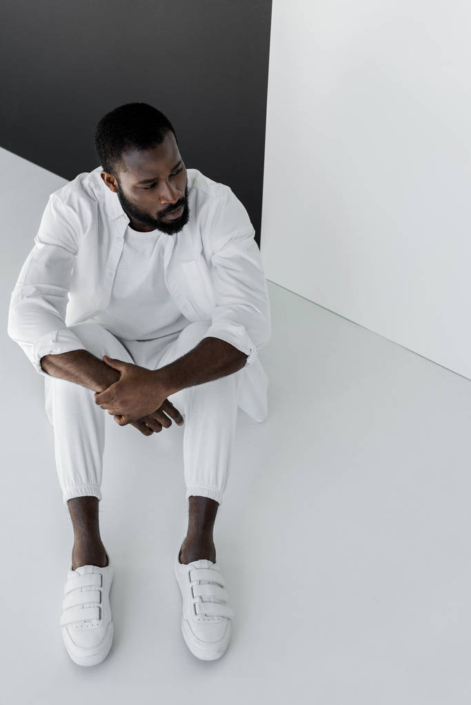 Hochwinkelaufnahme eines hübschen, stilvollen afrikanisch-amerikanischen Mannes, der in weißen Kleidern auf dem Boden sitzt - Foto, Bild