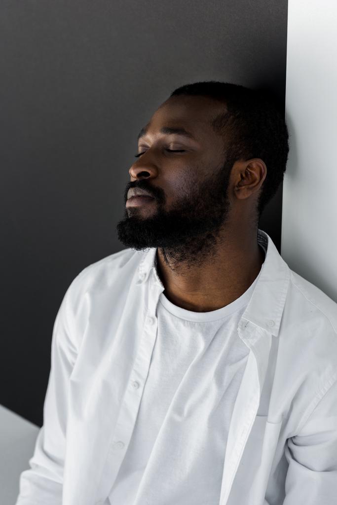 schöner stylischer afrikanisch-amerikanischer Mann in weißer Kleidung lehnt mit geschlossenen Augen an der Wand - Foto, Bild